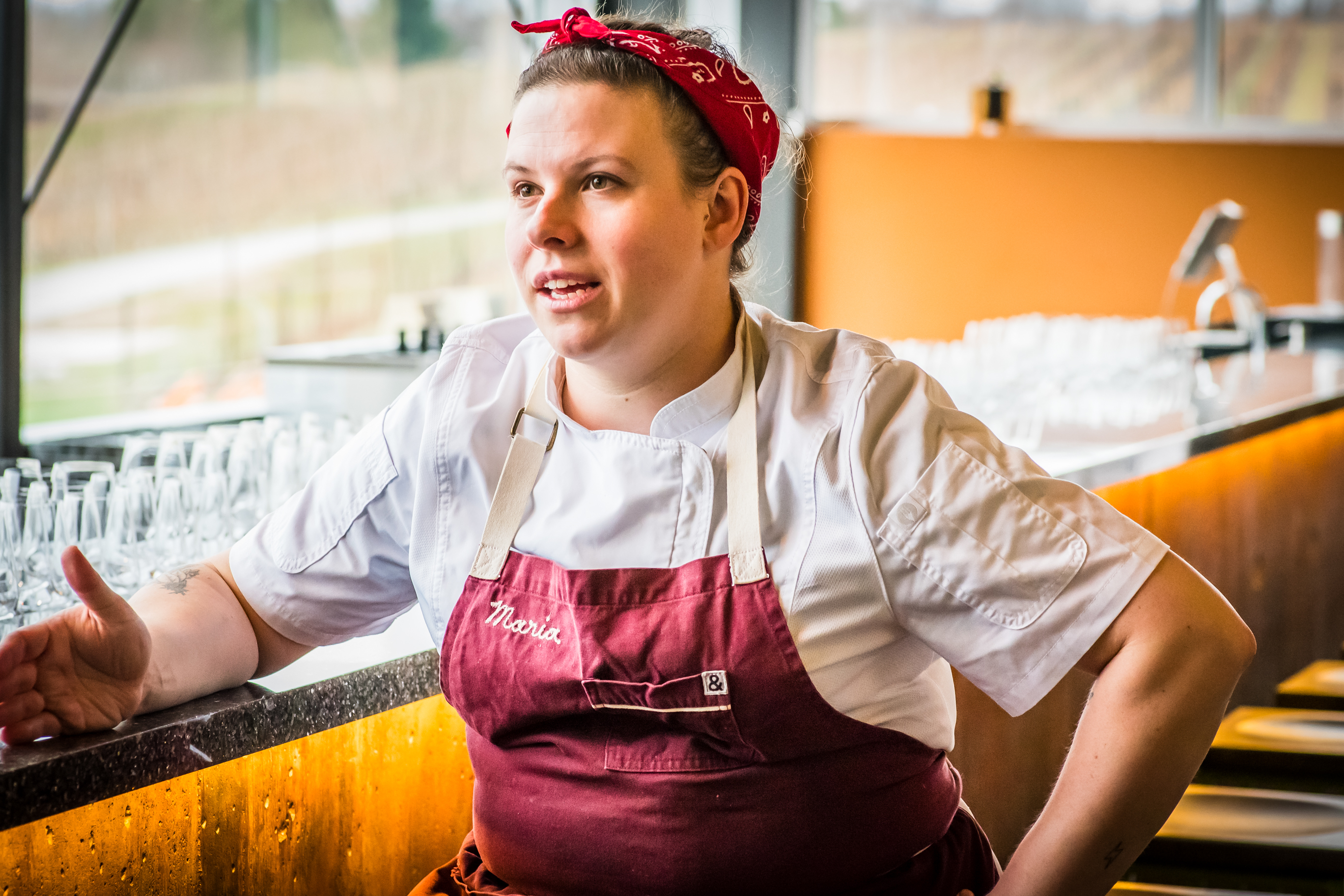 Maria in her chef jacket and burgundy apron, gesturing during a conversation at a kitchen counter